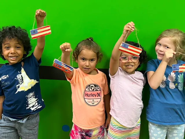 Four preschool children proudly raise handmade American flag ornaments while standing against a bright green wall.