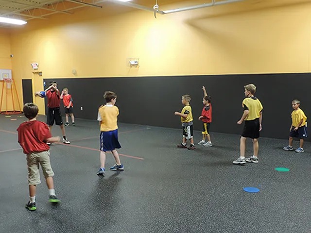 Elementary-age children in colored jerseys stand on a marked gym floor listening to a coach during an indoor physical education activity.