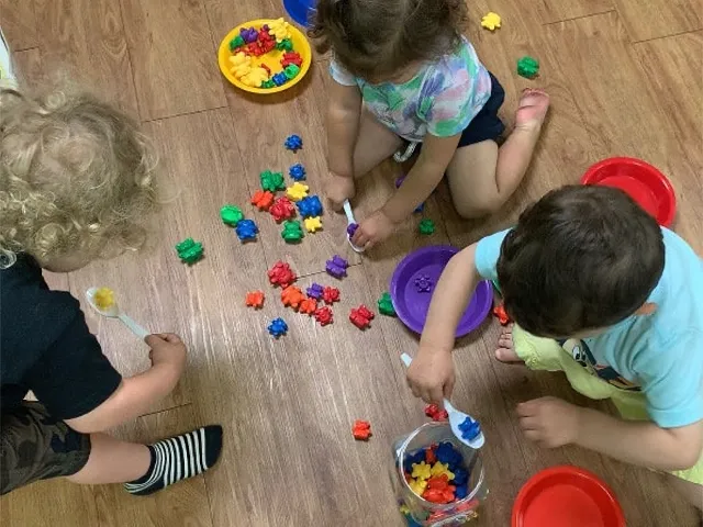 Three toddlers use plastic spoons to sort bright, multicolored counting bears into small round bowls on a wooden classroom floor.
