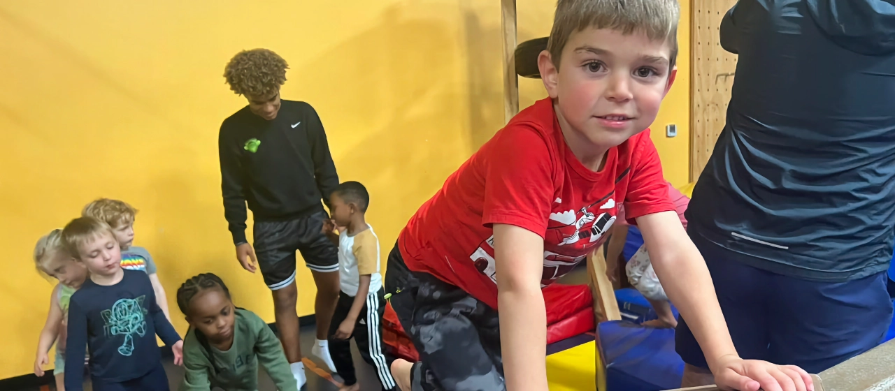 Young children practicing balance on a gymnastics beam in a gym with an instructor supervising.