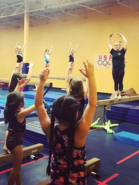 Children participating in a stretching exercise during a gymnastics class with their coach guiding them.